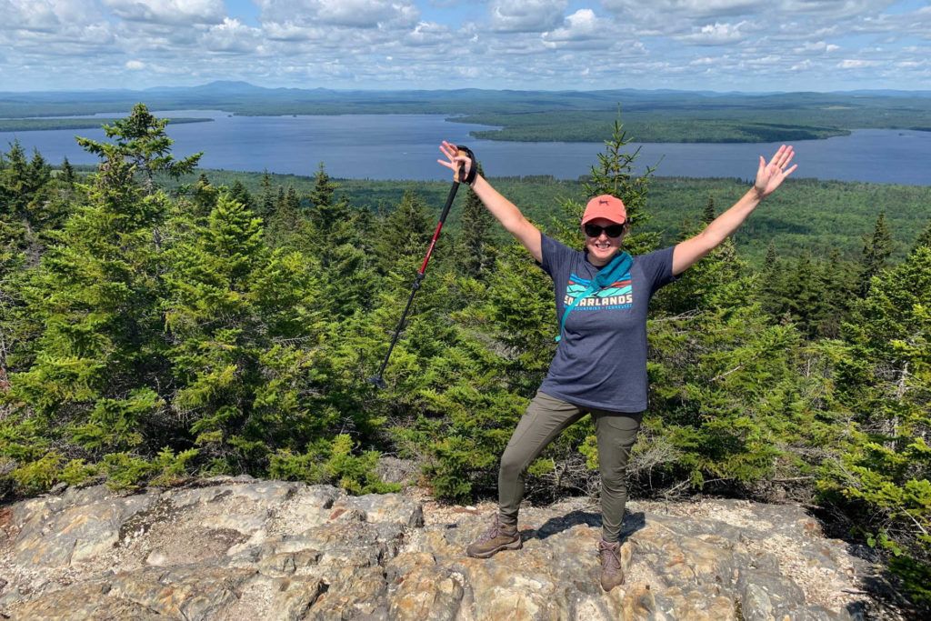 Hike with arms up holding a hiking pole at the top of Little Kineo Mountain in Maine