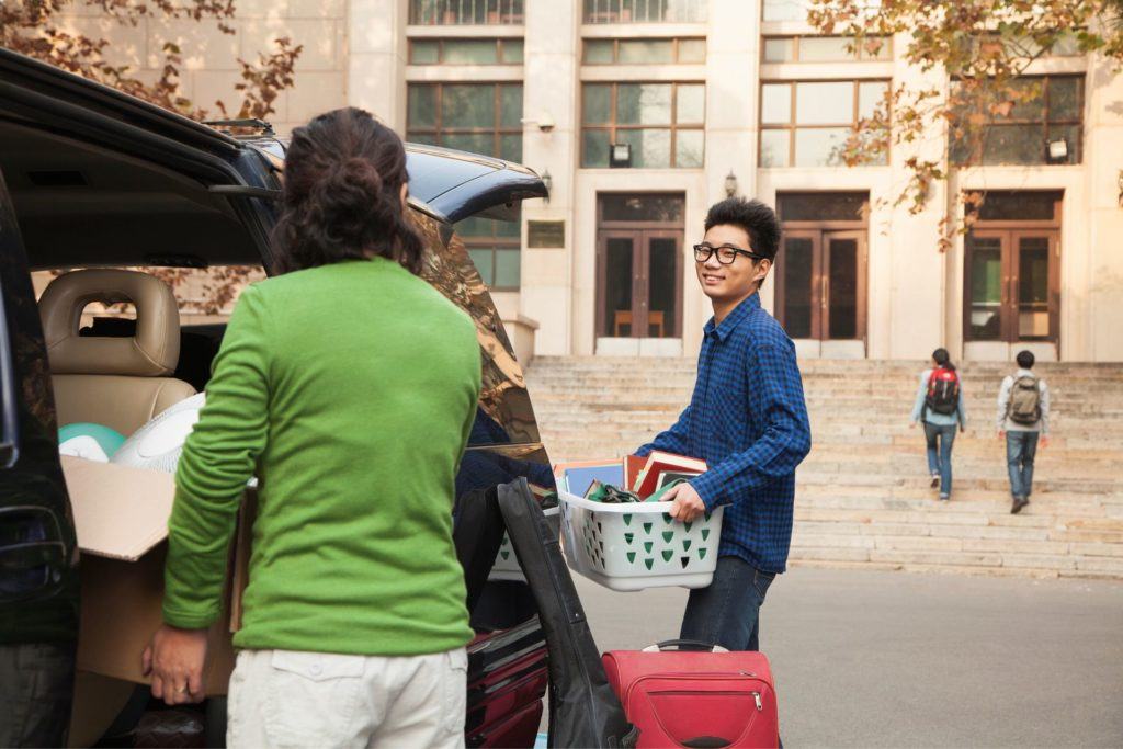 young Asian man carrying things into college dorm with mom in green sweater by car