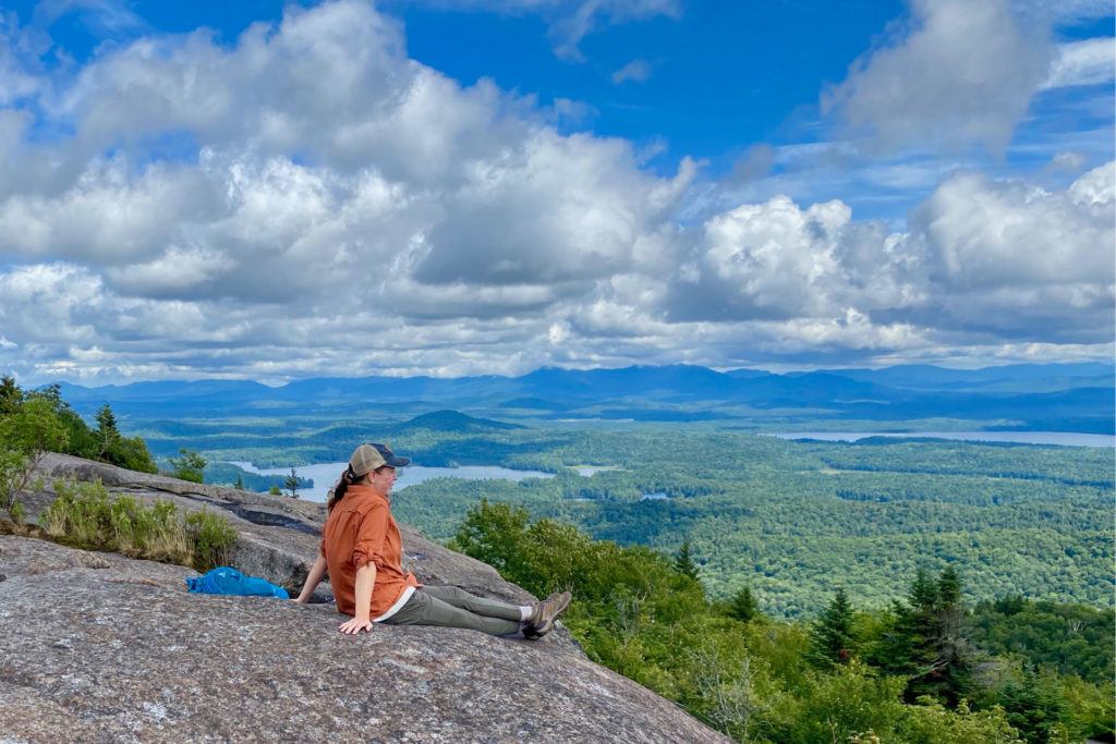 Female hiker sitting on rock summit looking out over lakes and valleys from St Regis Mountain in the Adirondacks
