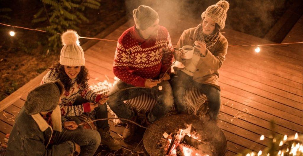 4 people sitting around a fire pit roasting marshmallows and drinking hot chocolate wearing sweaters and hats