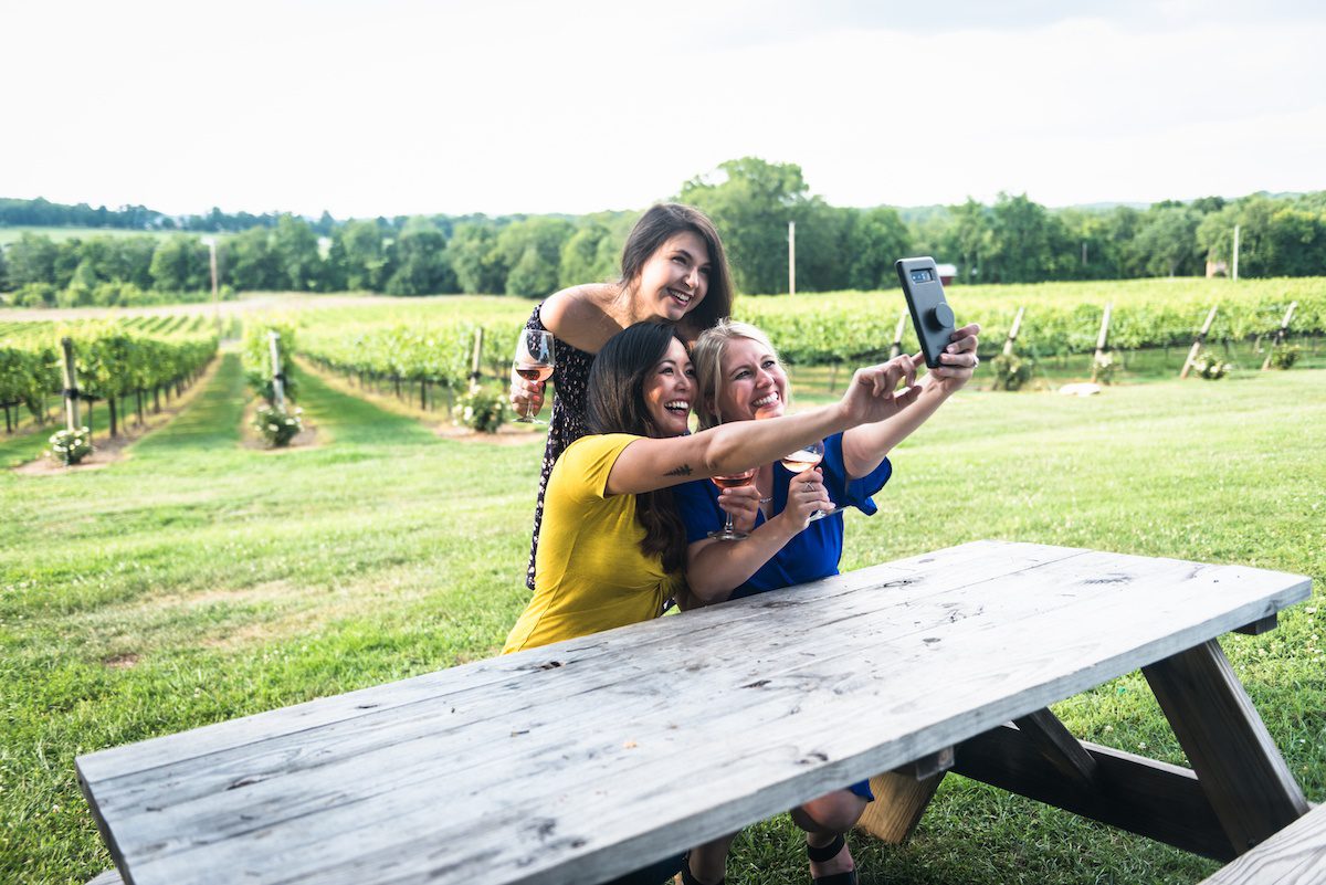 Three women holding wine at a picnic table in a vineyard taking a selfie with a phone