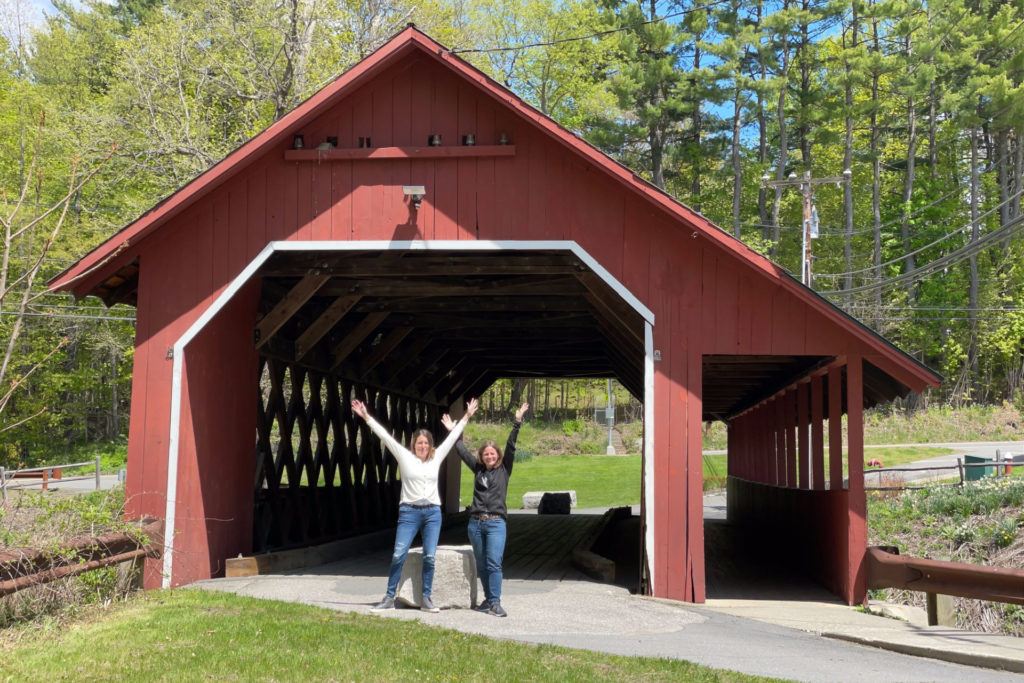 two women in front of Creamery Covered Bridge in Brattleboro Vermont with their arms raised