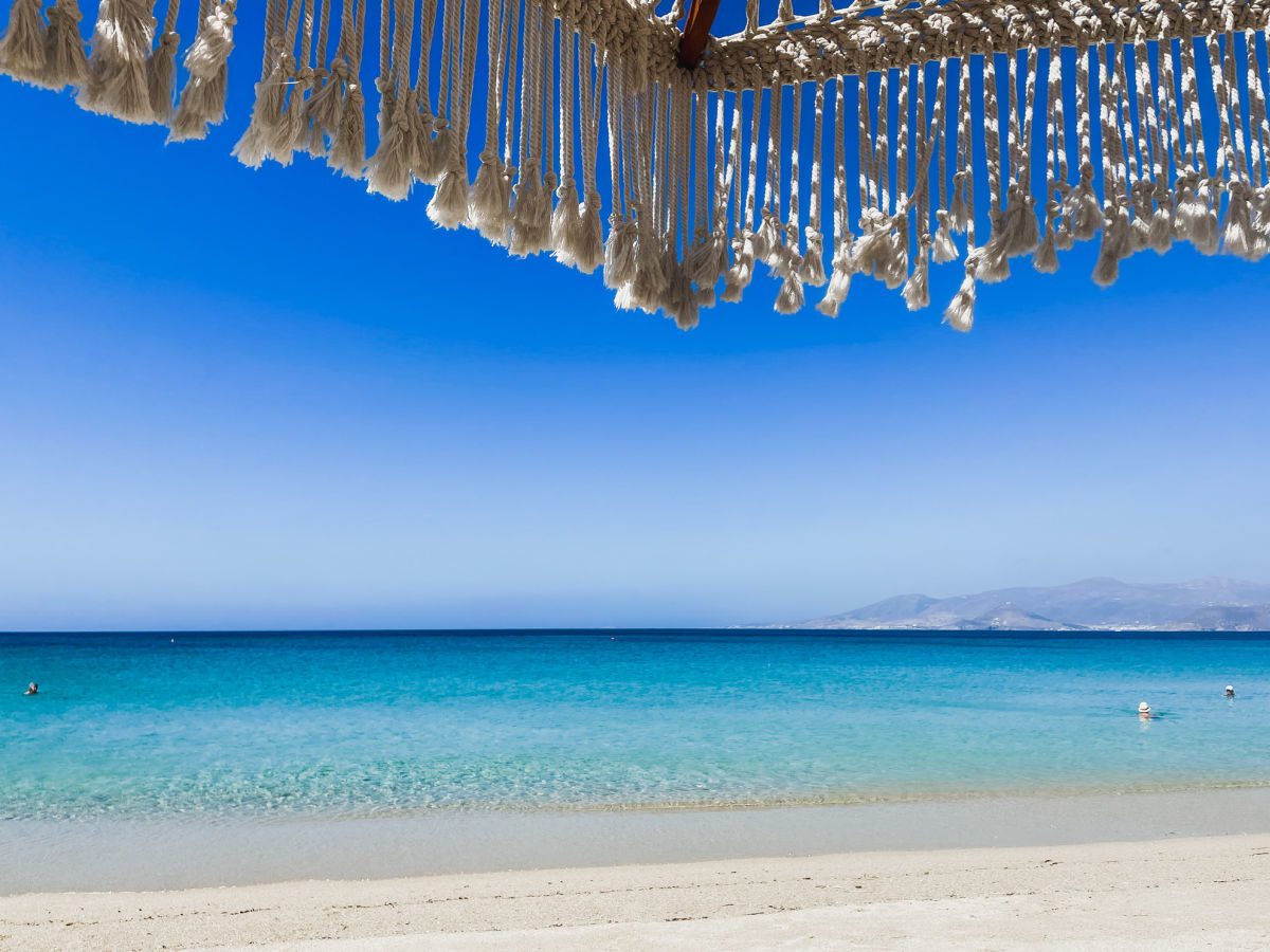 Agios Prokopios beach with blue water and white sand, looking out from under a rope umbrella at Paros in the distance