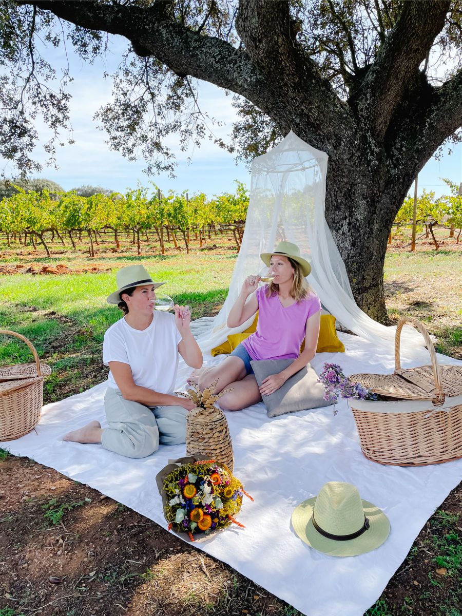 Two women sipping wine on a picnic blanket in a vineyard
