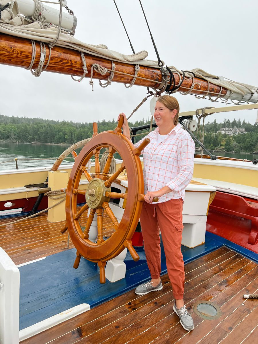 Tamara in orange joggers and a white and orange linen shirt at the helm of the Schooner Heritage