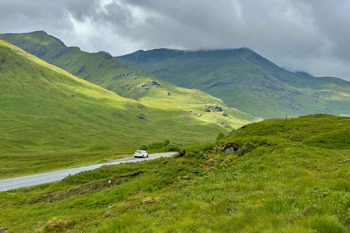 Road through Scottish Highlands with green hills and white car on the road